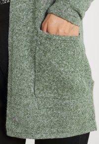 Ruka částečně zasunutá do přední kapsy zeleného pleteného kardiganu nošeného přes černé kalhoty.