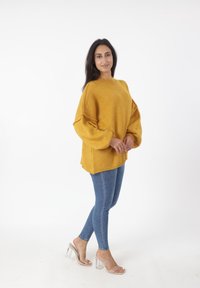 Maglione oversize color giallo senape con maniche a sbuffo, abbinato a jeans skinny blu e sandali con tacco trasparente, su uno sfondo bianco.