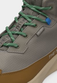 Γκρι και καφέ αθλητικό παπούτσι με πράσινα κορδόνια, γλώσσα από δίχτυ, μπλε θηλιά για τα κορδόνια και ορατή ετικέτα υφάσματος «DRYVENT».