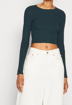 Vrouw draagt een donkergroene geribbelde crop top met lange mouwen en witte broek met hoge taille, waarbij haar buik en een deel van haar handen zichtbaar zijn.