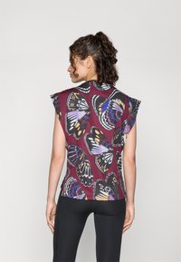 Blouse à manches courtes avec une base bordeaux ornée d'imprimés de papillons colorés. Texture lisse, coupe décontractée et design unique des épaules.