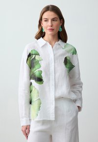 Camicia di lino bianca con grandi stampe floreali verdi e nere, chiusura con bottoni sul davanti e colletto. Tessuto liscio con maniche lunghe.