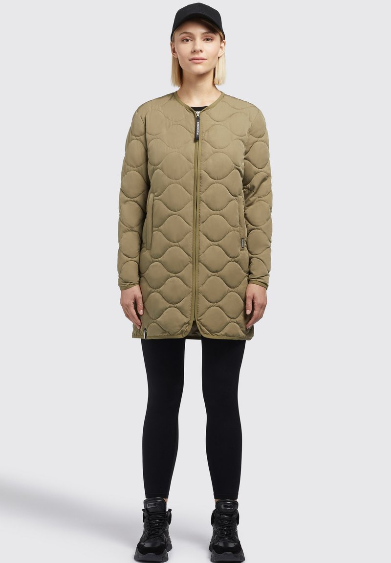 Steppjacke in olivgrün, lang geschnitten, mit einem Reißverschluss vorne, zwei Seitentaschen und rundem Ausschnitt, kombiniert mit schwarzen Leggings und Sneakern.