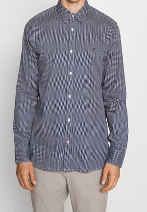 Marineblaues, gemustertes Hemd mit langen Ärmeln und Knöpfen, kombiniert mit hellbeigen Hosen, Modell von Hals bis Taille gezeigt.