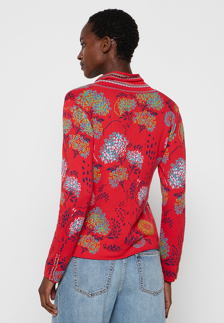 Czerwona bluzka z długim rękawem z kwiatowym wzorem w odcieniach niebieskiego, żółtego i białego. Dekolt ozdobiony szczegółowym wykończeniem; widoczna fakturowana tkanina.
