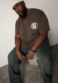 Brun bomulds t-shirt med et rundt logo og "S"-design på brystet, parret med blå denim jeans. Tilbehøret inkluderer sorte armbånd.