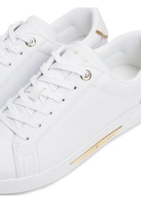 Bílé kožené tenisky s bílými tkaničkami, zlatými kovovými průchodkami a zlatým brandingem Tommy Hilfiger na boku a jazyku.