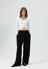 Wit geribd top met lange mouwen en een kruis-front ontwerp, gecombineerd met zwarte, wijde, hoge taillebroeken. Zwarte puntige schoenen maken de look compleet.