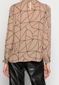 Blouse beige avec motif géométrique noir, manches longues, dos en goutte d'eau, tissu texturé et liens aux poignets, accompagnée d'un pantalon en cuir noir brillant.