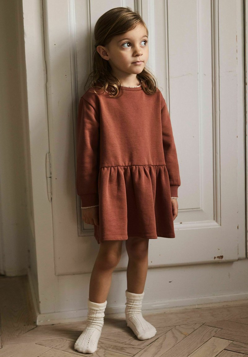 Jeune fille aux cheveux bruns portant une robe à manches longues couleur rouille et des chaussettes blanches tricotées, debout à l'intérieur près d'une porte blanche à panneaux.
