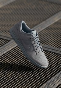 Grå Nike-sneaker med overdel i semsket skinn og lær, perforert tåboks, snøringer, polstret krage, og tykk gummisåle på en teksturert overflate.