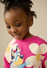 Felpa rosa acceso con grandi fiori bianchi e una grafica giocosa di un cane dei cartoni animati blu. Morbido cotone, scollo rotondo casual.