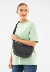 Borsa a tracolla in pelle nera con un design curvo e una texture liscia, abbinata a una maglietta verde chiaro e jeans blu.