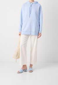 Hellblauer übergroßer Hoodie mit Fronttasche, kombiniert mit weißen, plissierten, weiten Hosenbeinen und blauen Open-Toe-Mules. Insgesamt glatte Textur.