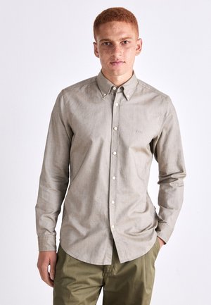 Camisa de botones color beige claro con mangas largas, textura sutil y un pequeño logotipo bordado en el pecho, combinada con pantalones verde oliva.