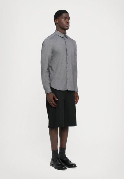 Uomo in piedi che indossa una camicia grigia a maniche lunghe con bottoni, pantaloni corti neri fino al ginocchio, calze scure e scarpe eleganti nere su uno sfondo bianco.