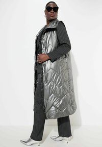 Silberne Weste aus Puffmaterial mit gestepptem Design und Druckknöpfen, über einem schwarzen Outfit getragen. Die Textur ist glänzend, mit hohem Kragen und offenen Seiten.