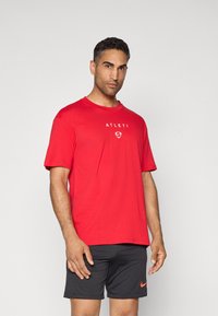 Rød bomulds T-shirt med korte ærmer og "ATLETI" logo i hvid, samt mørkegrå shorts med et orange Nike-logo.
