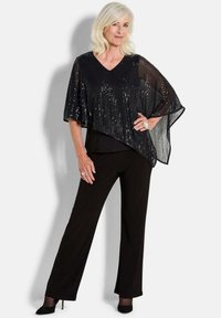 Schwarzer Pailletten-Poncho mit V-Ausschnitt-Design, über einem schwarzen Top getragen und mit schwarzen weiten Hosen kombiniert. Verfügt über eine strukturierte, schimmernde Oberfläche.