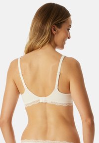 Reggiseno beige chiaro con rifiniture in pizzo. Presenta spalline regolabili, chiusura con gancetto e un finish in tessuto liscio. Vista dal retro visibile.