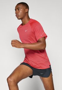Vörös sportpóló textúrált mintával, rövid ujjak és Nike logó. Fekete rövidnadrággal viselve, bemutatva a könnyű sportöltözet tervezését.