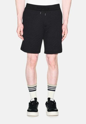 Uomo in piedi che indossa pantaloncini neri strutturati, calzini bianchi con strisce nere e beige e scarpe da ginnastica nere con suole bianche.