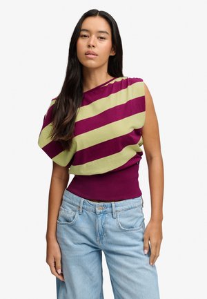Frau mit langen dunklen Haaren, die ein ärmelloses Oberteil mit breiten violetten und hellgrünen diagonalen Streifen sowie hellblaue Jeans trägt, steht vor weißem Hintergrund.