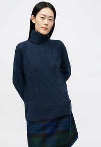 Pulover de culoare bleumarin cu guler tip tort de gât, cu un model din tricotaje cu cablu, purtat peste o fustă în carouri verzi și negre.