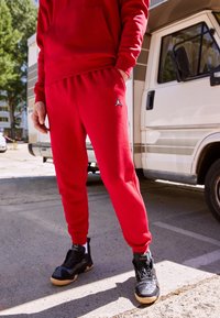 Červená mikina s kapucí a odpovídající tepláky vyrobené z měkké tkaniny, s vyšitým logem. Nošeno s černými teniskami a výrazným designem s vysokým profilem.