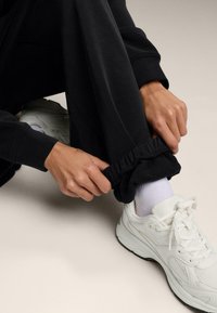 Čierne jogger nohavice so zúženými manžetami si prispôsobuje ruka, doplnené o biele tenisky a svetlé ponožky na neutrálnom pozadí.