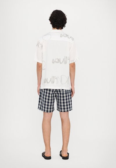 Short-sleeve white shirt with gray printed lettering, paired with navy and white checkered shorts. Footwear includes black sandals.