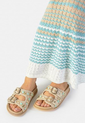 Pies con sandalias beige de punta abierta con hebillas decorativas y tachuelas turquesas, combinadas con una falda de punto a rayas azul, beige y blanca.