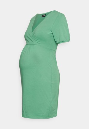 Vestido de maternidade verde feito de tecido suave, com decote em V, mangas curtas bufantes e uma silhueta ajustada que acomoda uma barriguinha em crescimento.