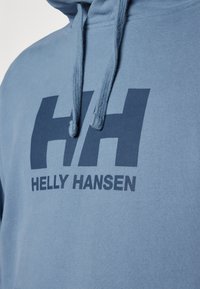 Felpa blu con cappuccio e cordini. Presenta un grande logo "HH" blu scuro e il testo "HELly HANSEN" sul davanti. Tessuto morbido.
