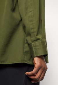 Camicia di cotone verde con maniche lunghe, caratterizzata da un polsino con dettaglio a bottone e un orlo dritto. Stoffa testurizzata, primo piano su un braccio.
