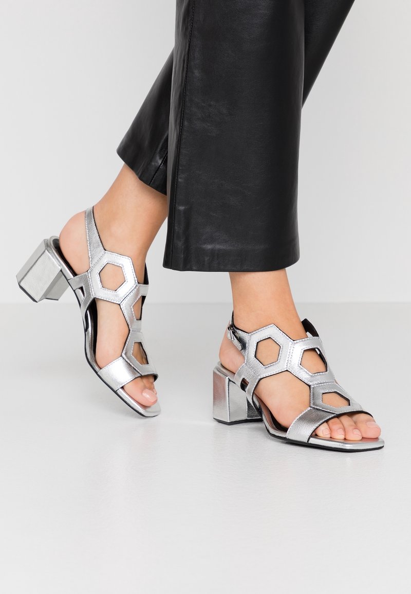 Pieds portant des sandales à talons blocs argentés à bout ouvert avec des découpes géométriques, associés à un pantalon en cuir noir raccourci, debout sur un sol clair.