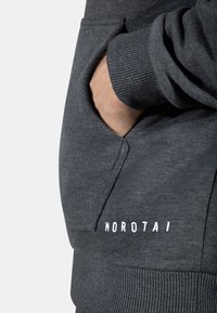 Tummanharmaa huppari, jossa on kengurupussi, ribbausta hihansuissa ja valkoiset bränditekstit "MOROTAI" vasemmalla alareunassa. Pehmeä kangas.