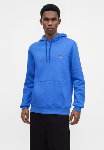 Homme portant un sweat à capuche bleu vif avec poche avant et un petit logo zèbre sur la poitrine gauche, assorti à un pantalon noir, debout devant un fond blanc.