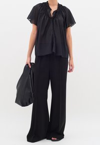 Femme portant un chemisier noir transparent à manches courtes amples et un pantalon noir à jambes larges, tenant un grand sac en cuir noir dans une main.
