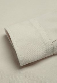 Tela texturizada color crema con puño doblado, que muestra costuras detalladas y patrones de tejido en una superficie textil suave.