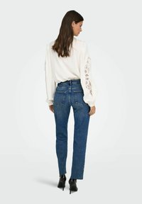 Blusa blanca con detalle de encaje en las mangas, combinada con jeans azul de cintura alta y tacones de aguja negros, vista desde la parte posterior.