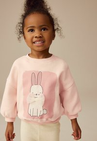Helles rosa Sweatshirt mit langen, puffigen Ärmeln und einem Grafikmotiv eines Kaninchen. Das Kaninchen hat gepunktete Ohren und eine Schleife, begleitet von einem Blumendetail.