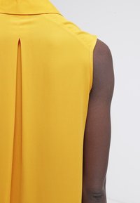 Bezramenná žlutá blůza s sklady na zádech. Tkanina je hladká s mírným leskem, což vytváří splývavý siluetu.