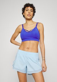 Sutiã de desporto roxo com alças cruzadas, combinado com calções azul claro com cintura elástica e bolsos laterais. Tecido liso e elástico.