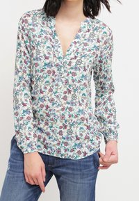 Blouse à manches longues à motif floral avec un col en V, présentant un fond blanc, des fleurs colorées en rose et bleu, et des boutons décoratifs au niveau du cou.
