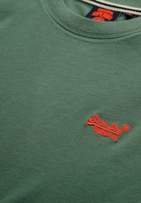 Sudadera verde con cuello redondo y pequeño logo rojo de SuperDry bordado en el pecho, además de una etiqueta de SuperDry en negro y naranja dentro del cuello.