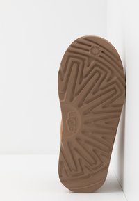 Suela de un zapato UGG marrón, que presenta un patrón de bandas en zigzag y un logo circular, con una superficie texturizada y acentos elevados alrededor de los bordes.