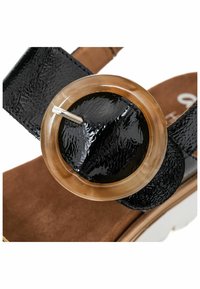 Sandale en vernis noir avec un accent circulaire en ambre et une bride texturée. Semelle en suède marron avec une semelle de couleur claire.