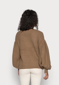 Pull en tricot marron avec des manches bouffantes et une texture côtelée. Il présente un col rond et une coupe décontractée, associé à un pantalon de couleur claire.