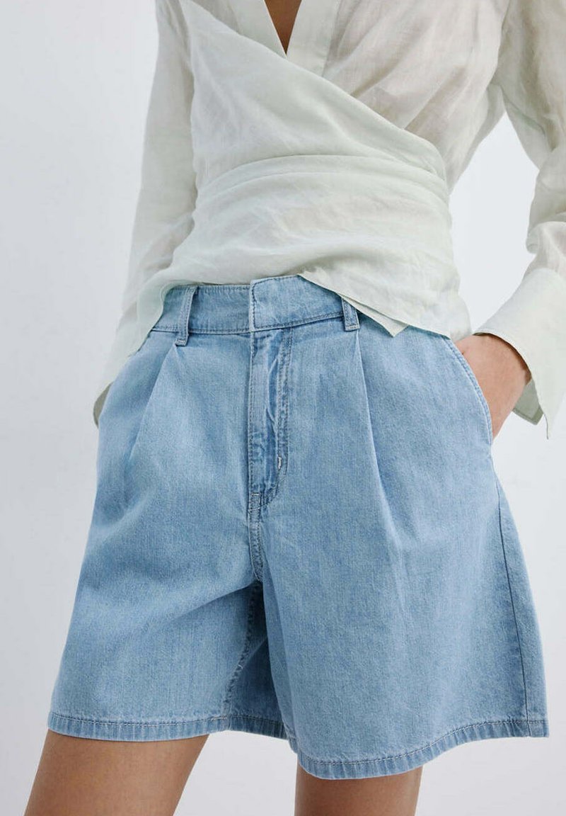 Person trägt hellblaue Jeans-Shorts und ein weißes Wickel-Oberteil mit langen Ärmeln, eine Hand in der Tasche.